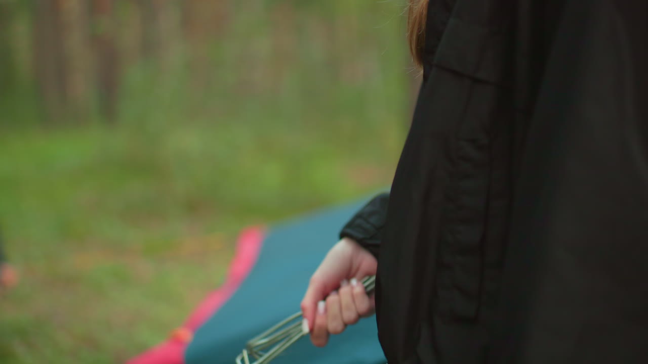 Close up of lady jiggling metal pegs in hand while standing near outdoor camping tent in forest, with soft background blur, casual clothing, and natural greenery