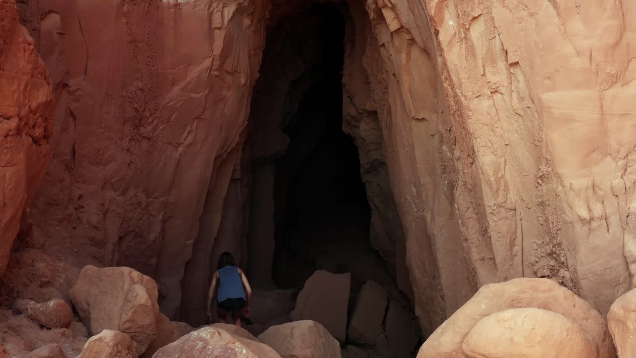 dron aéreo dolly en la toma de una joven delgada de cabello castaño bajando rocas rojas y caminando hacia la gran entrada de la cueva de la guarida del duende dentro del parque estatal de utah, valle del duende
