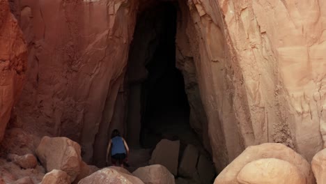 dron aéreo dolly en la toma de una joven delgada de cabello castaño bajando rocas rojas y caminando hacia la gran entrada de la cueva de la guarida del duende dentro del parque estatal de utah, valle del duende