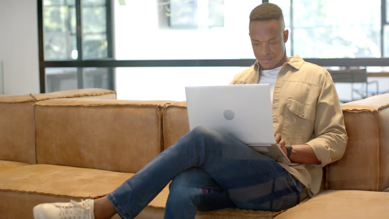 Working on laptop, African American man sitting on couch in modern office environment