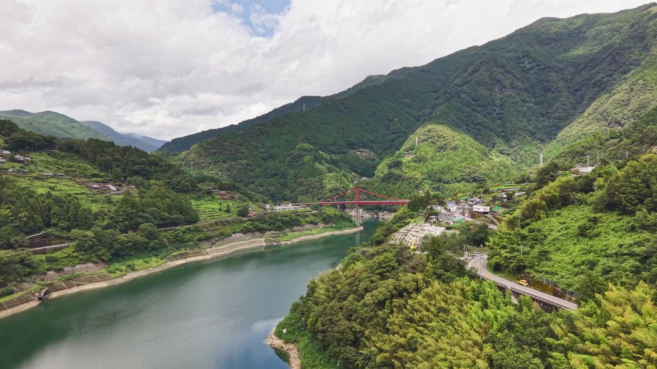 Cảnh quay drone bay về phía một cây cầu đỏ nổi bật bắc qua con sông xanh, nằm giữa những ngọn núi phủ rừng và một ngôi làng núi Nhật Bản đẹp như tranh vẽ
