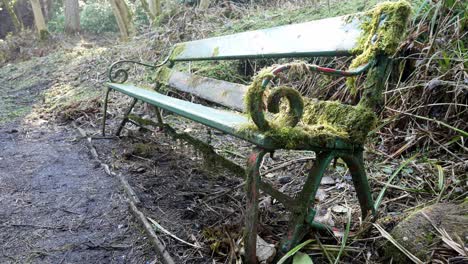 mossy neglected weathered green curled wrought iron wooden bench abandoned in woodland forest