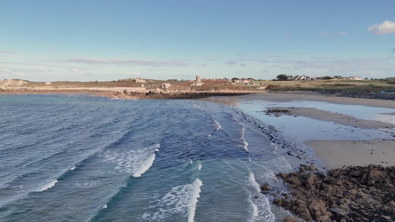 Lancresse Bay Guernsey: Flyv over kystlinjen med gylden sandstrand og klippefremspring på en solskinsdag med roligt, klukkende vand og flyvning mod Martello Tower på den fjerne side af bugten