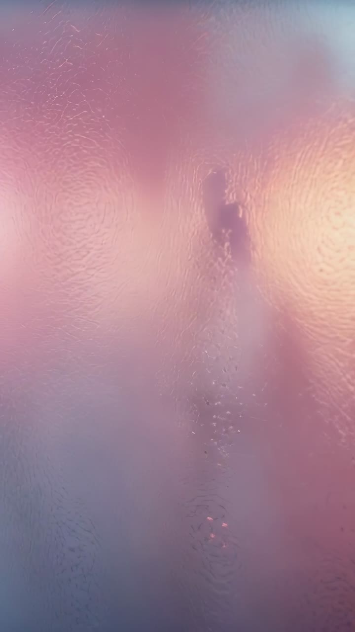 Vertical video: Droplets streaming down shower glass revealing silhouette emerging in warm glow
