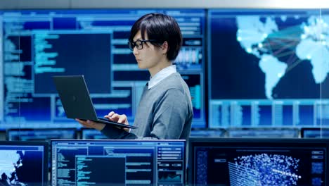 in the system control room it technician holds and works on a laptop, in background multiple displays with graphics. facility works on artificial intelligence, big data mining, neural network, surveillance project.