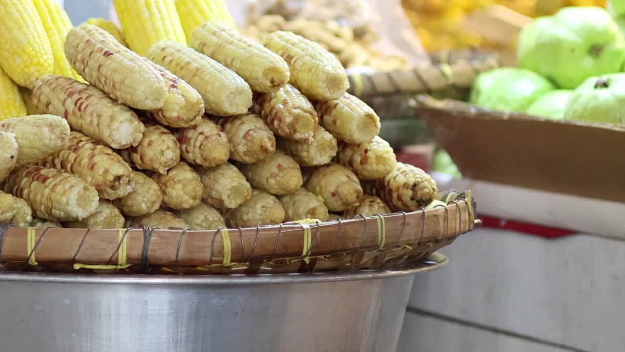 A vibrant market scene featuring neatly stacked corn cobs and potatoes in a large metal bowl.