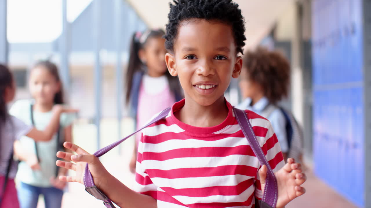 Smiling boy in striped shirt holding backpack straps at school with friends