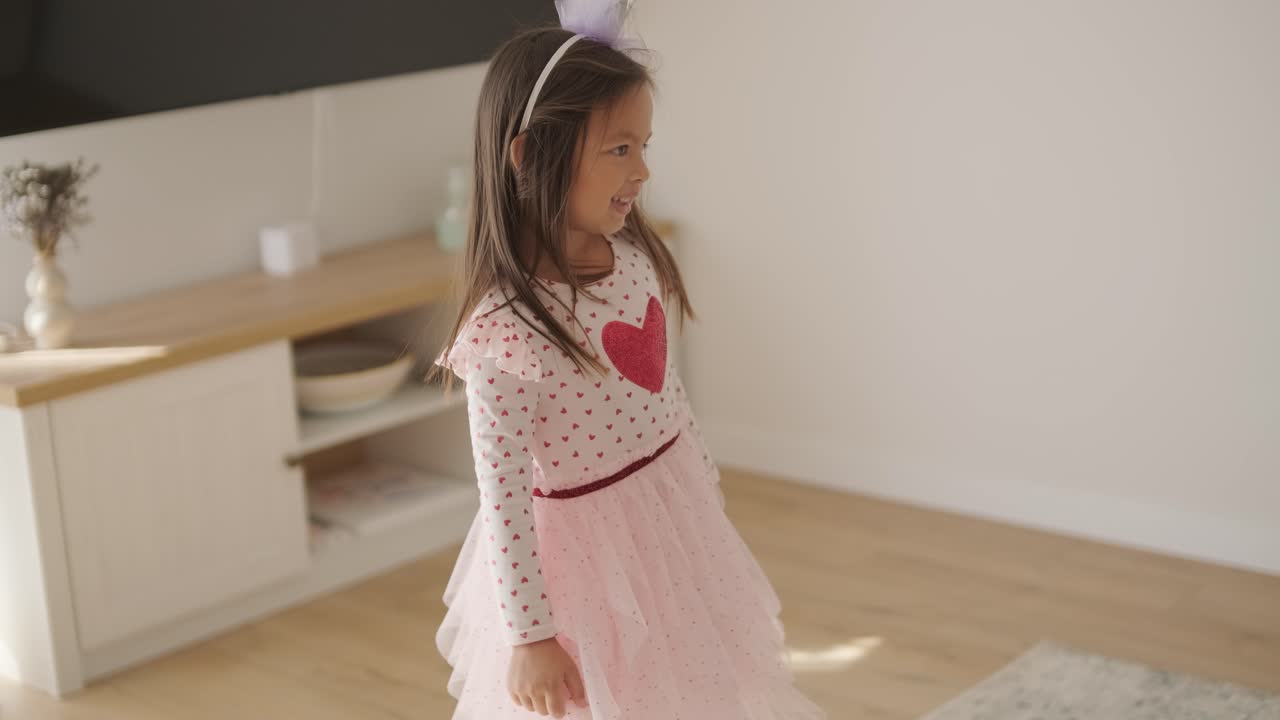 молодая девушка весело танцует в светлой, уютной гостиной. она носит розовое платье с дизайном сердца, играюще движется в солнечном, минималистском пространстве с нейтральным декором и мягким естественным светом.