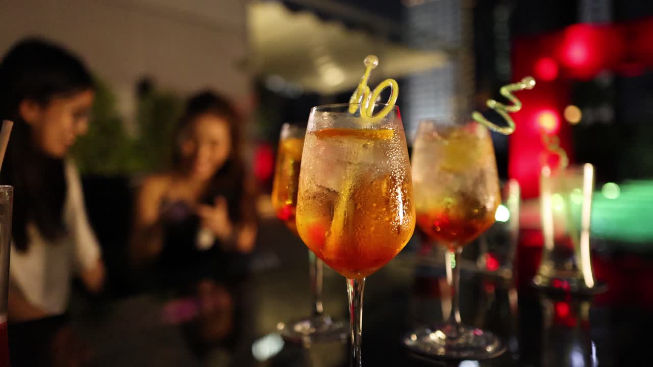 Two women enjoy lively conversation at a night party, with vibrant cocktails and decorative lighting in a shallow focus, festive atmosphere