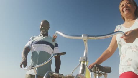 una pareja de ancianos afroamericanos sonrientes caminando con bicicletas en una playa soleada