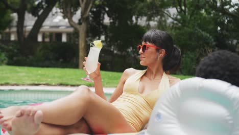 Happy-diverse-couple-wearing-swimming-suits-with-inflatables-and-drink-at-swimming-pool-in-garden