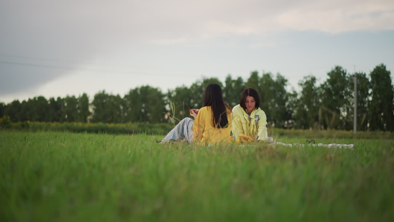 한 커플이 초원에서 석양을 바라보며 피크닉을 즐기고 있어요. 담요에 기대어 앉아 수평선을 바라보며 조용히 대화를 나누고 있으며, 따뜻한 햇살이 비추고 캐주얼한 데님 의상을 입고 편안한 분위기예요. 멀리 보이는 나무들이 장면을 감싸고 있어요.