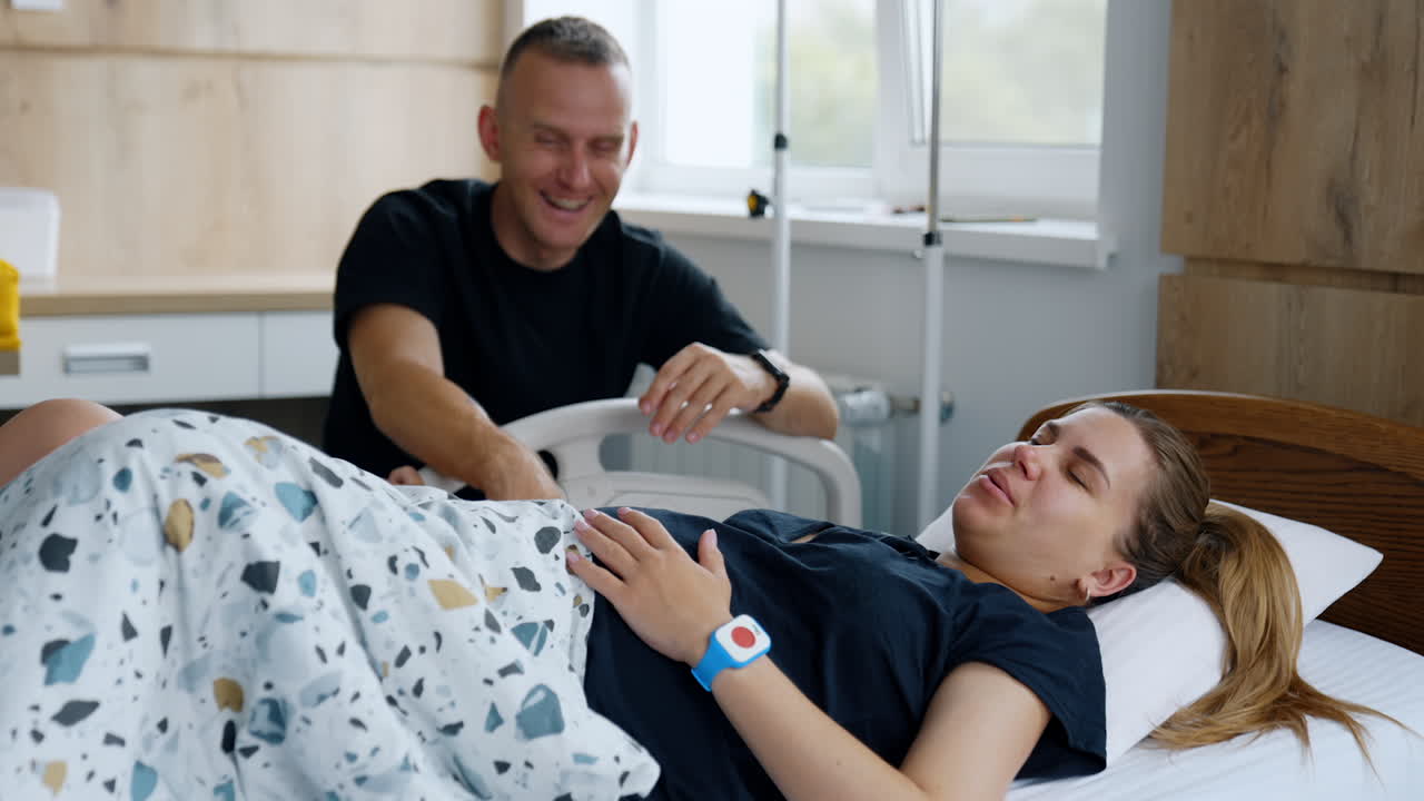 Man in black shirt sits near the bed with his wife. Woman smiles and then feels pain from the contractions. Childbirth concept.
