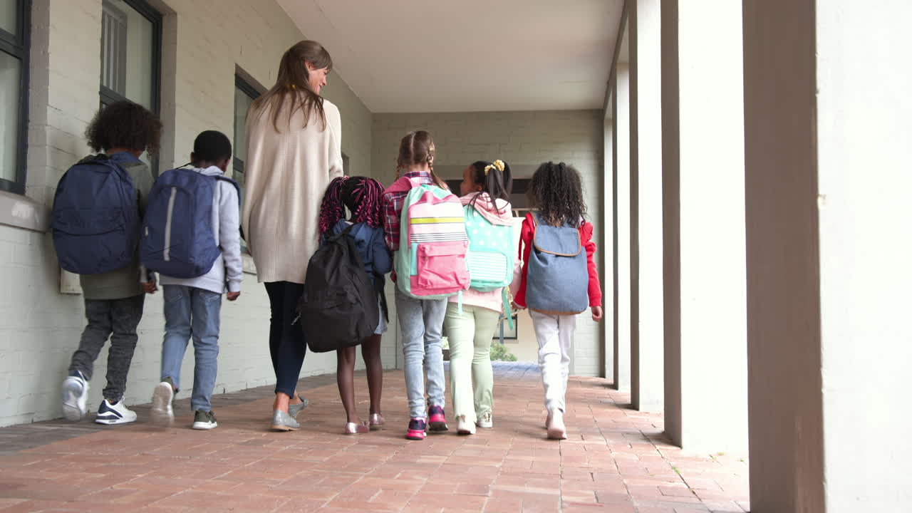 Teacher walking with diverse group of children wearing backpacks at school