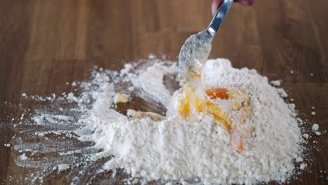Person-mixing-with-fork-an-egg-with-wheat-flour-on-table-to-make-homemade-food