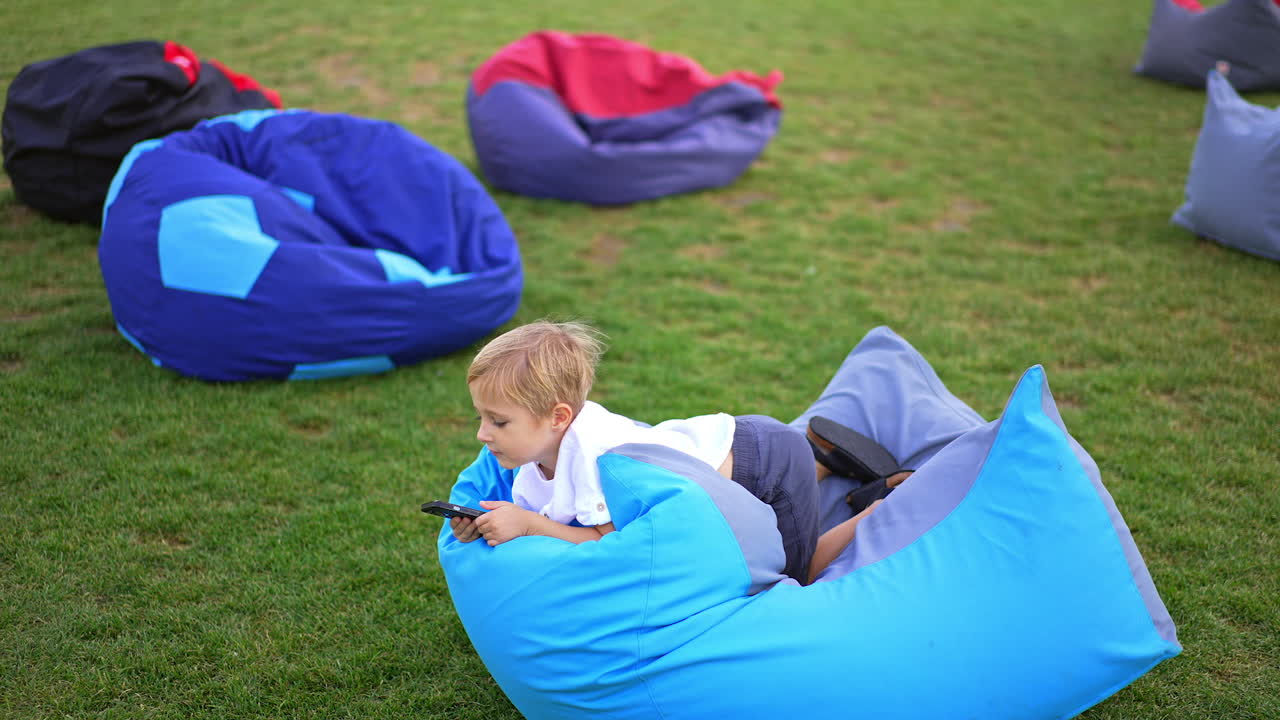 Leisure time spent with gadgets outdoors. Blond kid in a comfortable bag chair using smartphone. Green grass with bean bag chairs at backdrop.