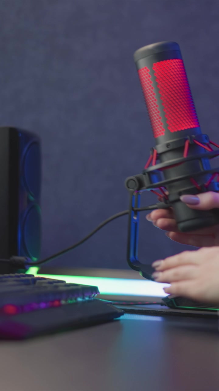 Una mano si allunga verso un microfono rosso e nero con illuminazione LED, poi lo regola vicino alla base. La scena include mani su una tastiera RGB sotto una luce ambientale soffusa, su sfondo scuro