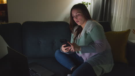 woman sitting on sofa at home at night streaming or watching movie or show on laptop and scrolling internet on phone 11