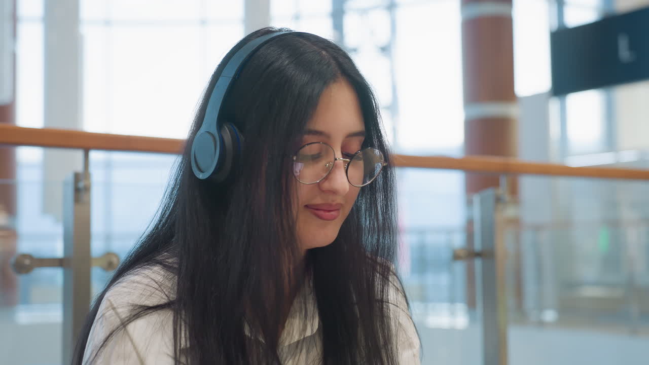 Portrait of young woman with long dark hair wearing glasses and headphones smiling gently while gazing upward with soft natural light and modern interior elements in background