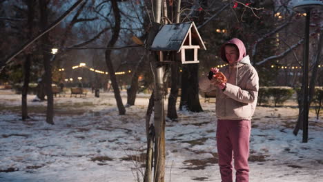 mujer joven de pie en el parque de invierno, dejando caer comida en la casa de pájaros para pájaros con tierra cubierta de nieve y luces del parque iluminadas