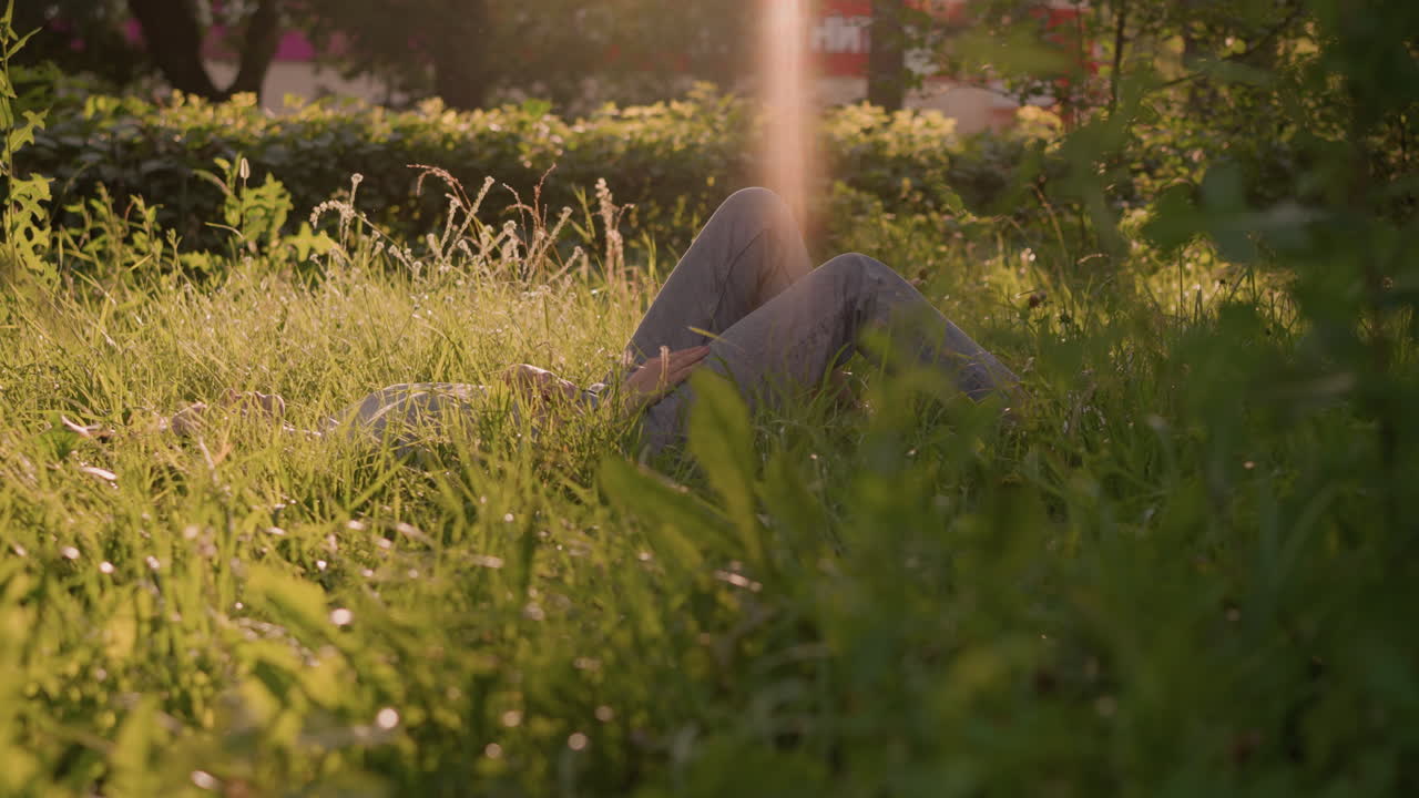 человек лежит на травяном поле под теплым солнечным светом, окруженный высокой травой и естественной зеленью, ноги согнуты, руки опираются на тело, на заднем плане деревья и кустарники