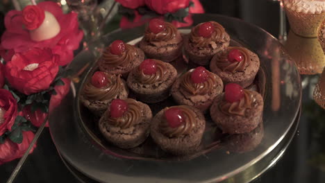 several chocolate cupcakes with generous topping of dulce de leche and a candied cherry on top, served on a silver tray during a party with other sweets and flowers in the background, slow moton shot