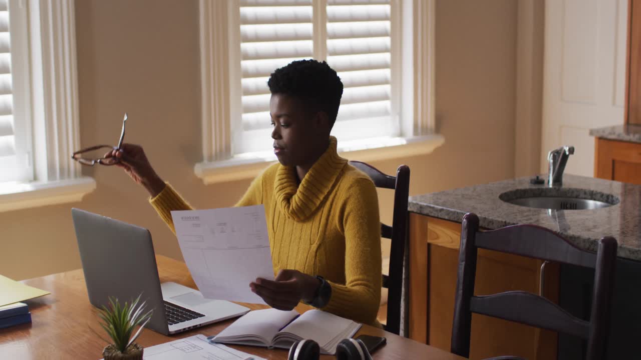 African american woman holding a document and using laptop while working from home