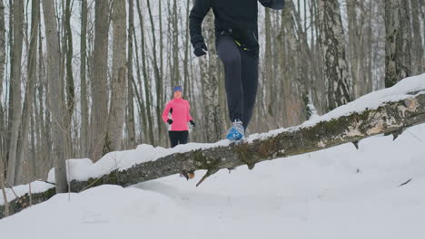 a man and a woman run in the park in winter and jump over a fallen tree. step over the obstacle