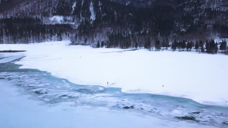 Two-people-are-standing-in-a-big-snow-field-in-a-valley-with-a-icey-lake