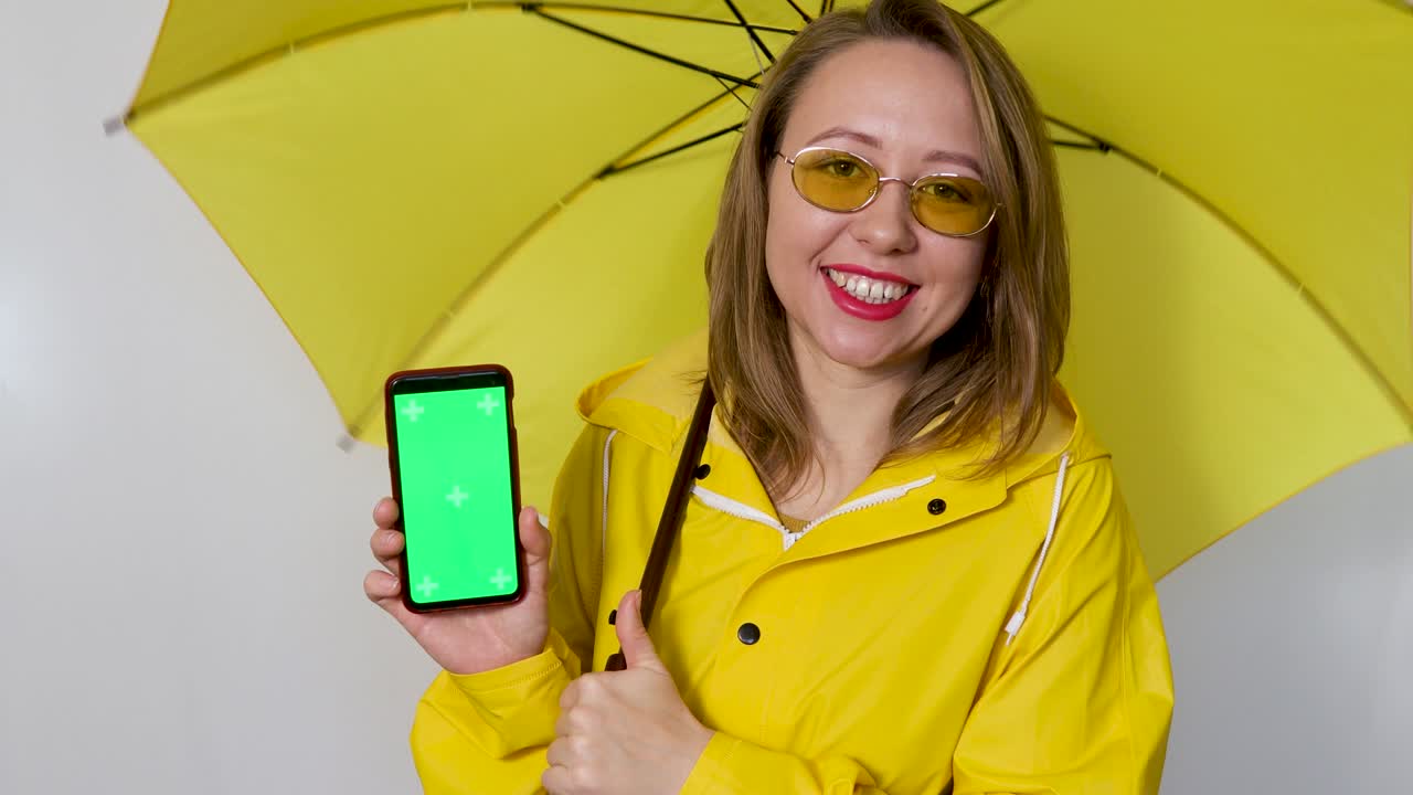 노란색 레인코트를 입고 우산을 쓴 소녀가 시청자에게 녹색 화면을 보여줍니다. 색 배경에 고립되어 있습니다.