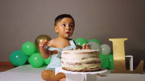 lindo bebé latino celebrando su primer cumpleaños mordiendo y rompiendo un pastel con globos verdes en el fondo