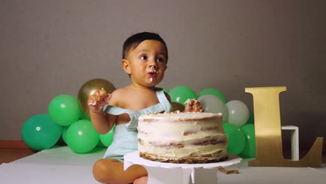 cute latin baby toddler celebrating his 1st year birthday biting and smashing a cake with green balloons in the background