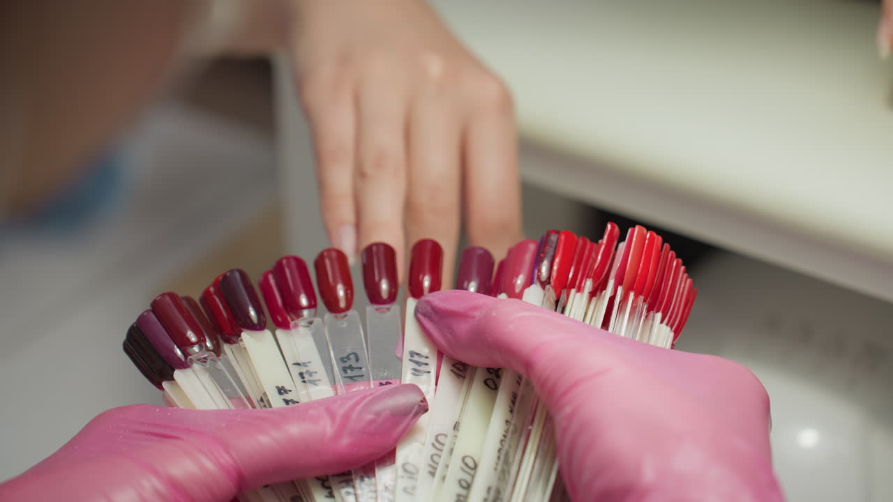 Nail technician in pink gloves holding nail polish color samples in shades of red and burgundy while customer hand rests nearby, evaluating choices during manicure session inside well-lit nail salon
