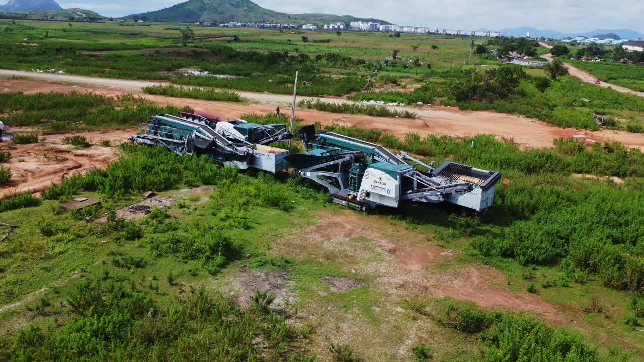High angle view of a large mobile crusher standing on lush, green farmlands in Nigeria, Africa on a sunny day. A wheelbarrow pusher and labourer is seen behind