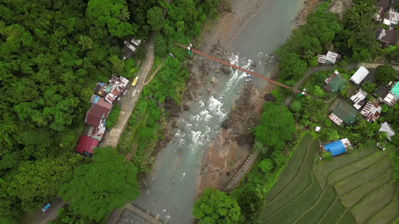 A wide aerial angle settles over a forested valley with a rocky river and red bridge centered in frame. Roads and rooftops flank both banks as cultivated fields stretch into the layered terrain