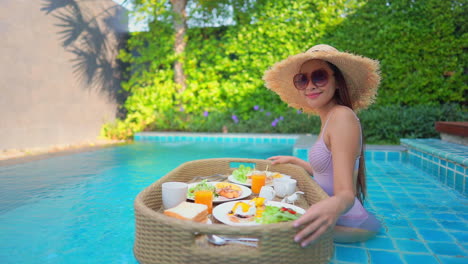 vista lateral de un turista asiático sentado en la piscina con una bandeja de desayuno flotante llena de comida deliciosa y saludable