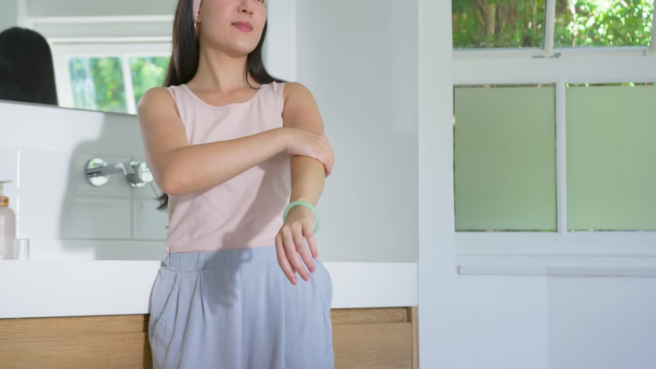 Asian woman wearing pink tank top adjusting green bangle rubbing arm placing headband in bathroom