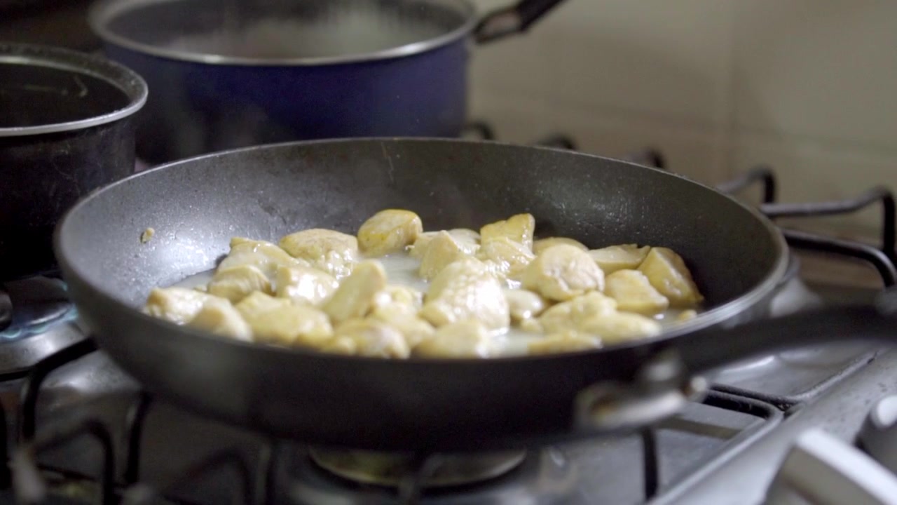 Pezzi di pollo che friggono in padella su fornello a gas durante la preparazione di un pasto fatto in casa, cuocendo a fuoco costante mentre sviluppano un colore dorato e una consistenza ricca