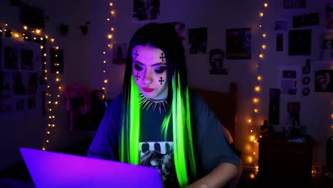 woman with glowing green hair and goth makeup working on a laptop in a dimly lit bedroom.