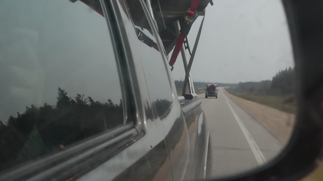 Filmagem manual enquanto se dirige no norte de Manitoba, Canadá, câmara focada no espelho lateral. Canoa na caminhonete e outro veículo com duas canoas atrás, estrada visível no reflexo, mão segurando a câmara.