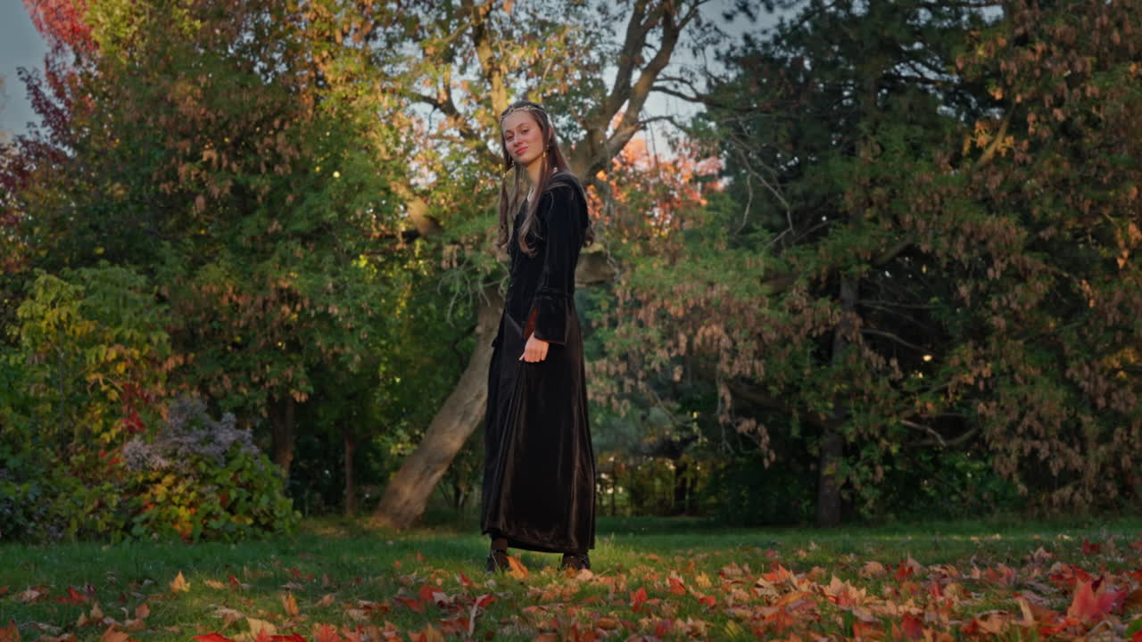 Ripresa cinematografica con carrello a destra di una donna medievale in abito di velluto nero, in piedi con grazia e con aria serena in una foresta autunnale naturale, con calda luce solare e foglie colorate cadute