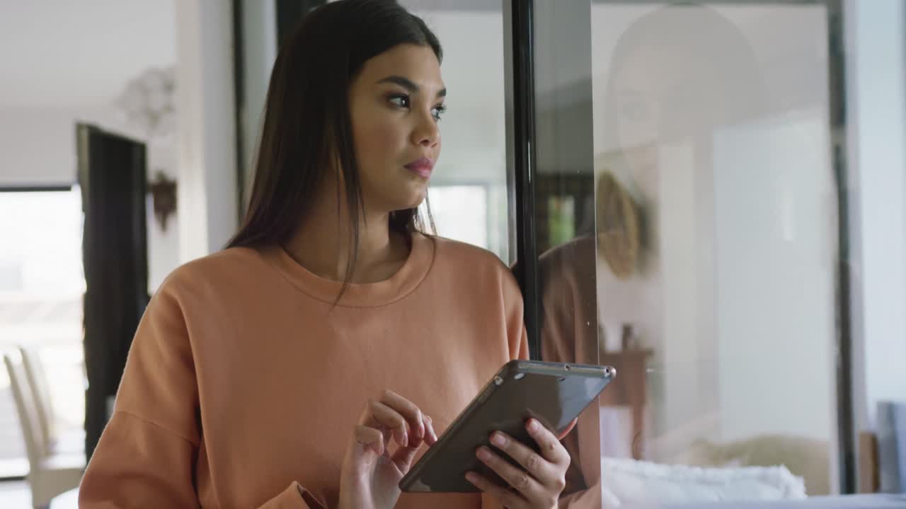 Video of thoughtful biracial woman standing at window and using tablet