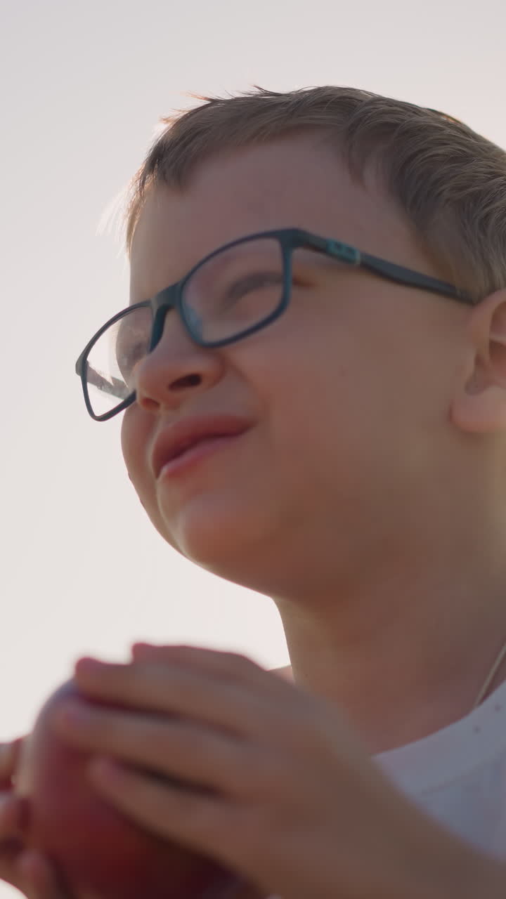 Close-up shot of a young boy wearing a white shirt and glasses, laughing as he takes a bite of a red apple in a sunlit field. The image captures a moment of pure joy and connection with nature