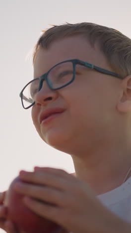 close-up shot of a young boy wearing a white shirt and glasses, laughing as he takes a bite of a red apple in a sunlit field. the image captures a moment of pure joy and connection with nature