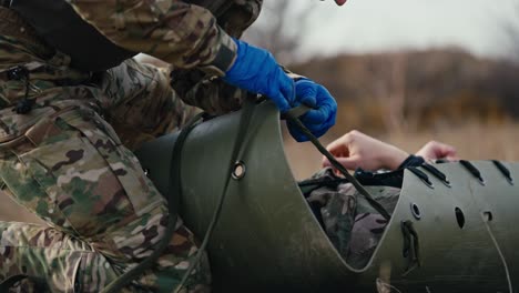 Desde-Un-Costado,-Un-Médico-Militar-Confiado,-Con-Ropa-Verde-De-Camuflaje-Y-Guantes-De-Goma-Azules,-Ata-Correas-A-Una-Camilla-Para-Trasladar-A-Un-Soldado-Herido-Durante-Operaciones-De-Combate-En-La-Estepa,-En-Las-Afueras-De-La-Ciudad.