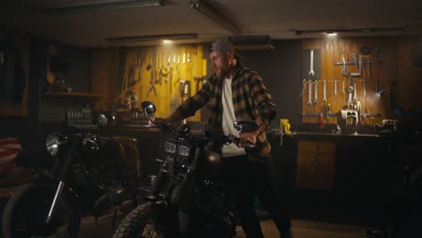 A-confident-guy-in-a-checkered-shirt-brings-in-a-motorcycle-and-prepares-for-repairs-in-the-workshop