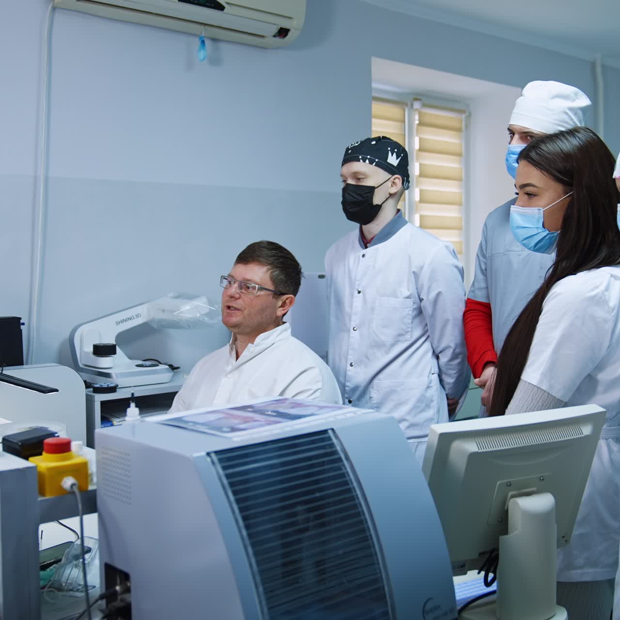 Un grupo de estudiantes con máscaras de pie detrás del profesor mira la pantalla del ordenador. Un médico muestra la radiografía de la dentadura postiza a futuros dentistas.