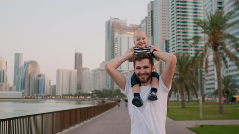 Ein-Junger-Vater-In-Einem-Weißen-T-Shirt-Und-Einem-Hipster-Mit-Borsten-Geht-Mit-Seinem-Sohn,-Der-Auf-Seinem-Hals-Sitzt,-An-Der-Uferpromenade-Vor-Dem-Hintergrund-Der-Modernen-Stadt-Im-Sommer-An-Der-Uferpromenade-Spazieren