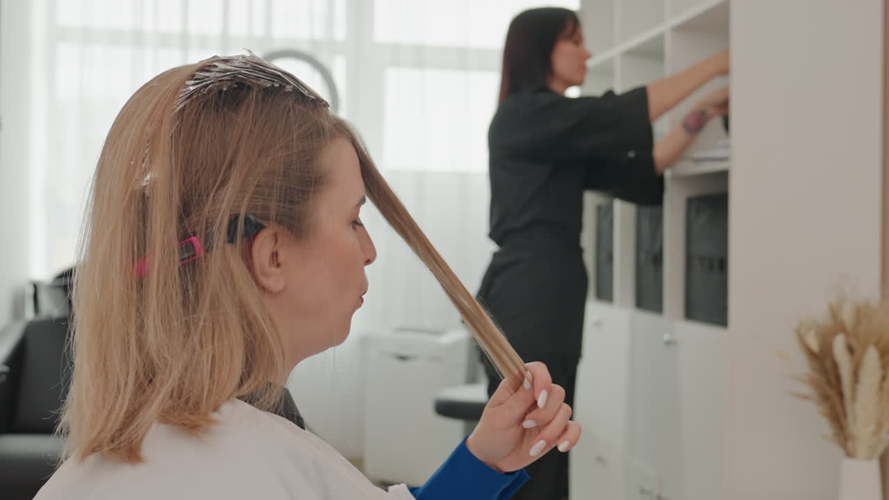 Blonde woman in salon sitting, twirling highlighted strand while stylist reaches cabinet to prepare color tools, foils visible in hair, bright modern studio with shelves, products and dried vase