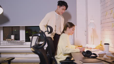 Male-Employee-Working-On-Laptop-Computer-And-Then-Approaching-His-Female-Colleague-To-Ask-Her-Something